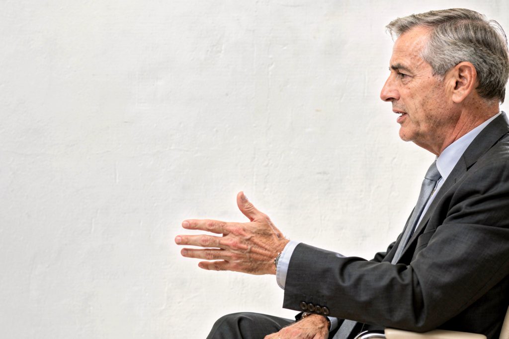 An older man in a suit gestures expressively with his right hand, seated against a plain white background, suggesting a serious and engaged conversation.