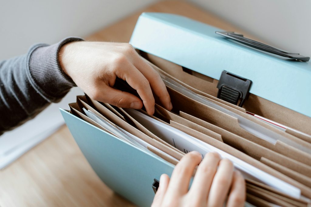 A person organizes documents in a turquoise accordion folder on a wooden desk, conveying a sense of neatness and attention to detail.