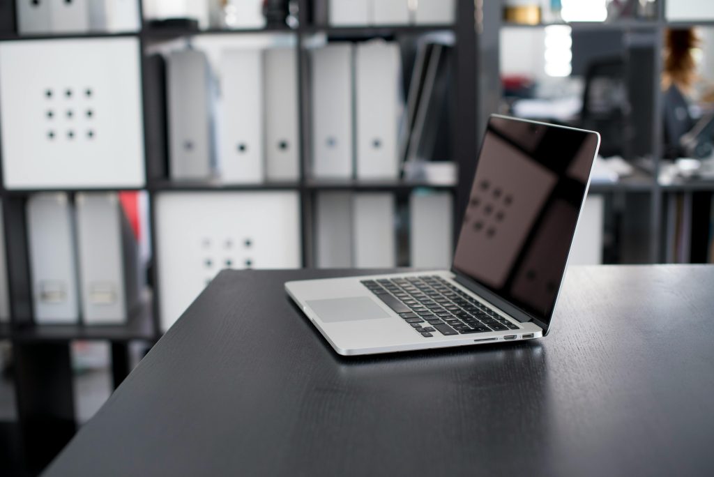 Open laptop on a black desk in a modern office. Background features shelves with white and black binders, suggesting organization and productivity.