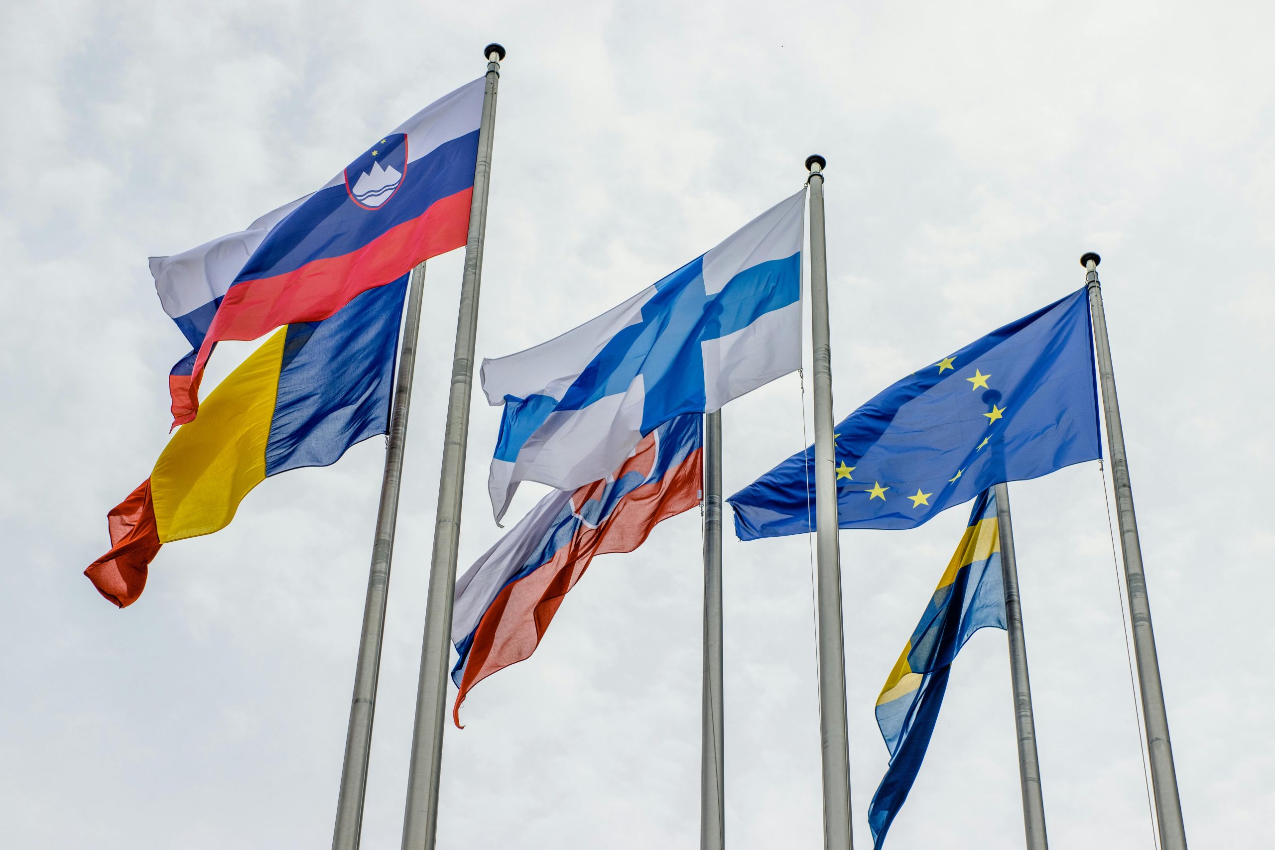 Five flags on tall poles wave against a cloudy sky. The flags include Slovenia, EU, and others, symbolizing unity and international cooperation.