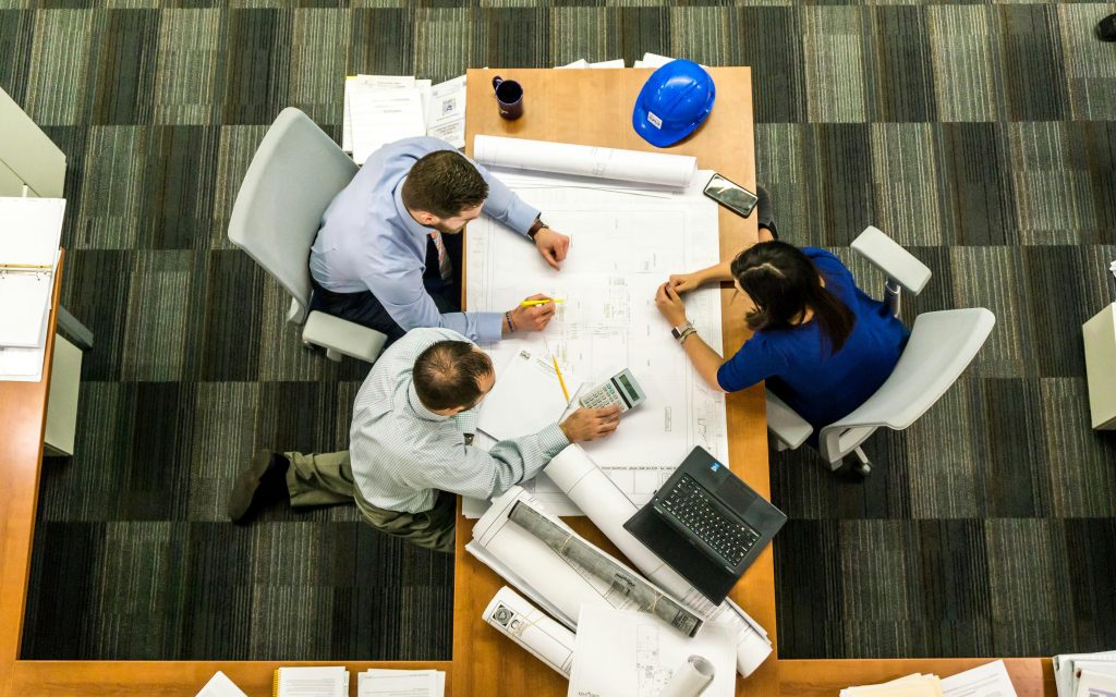 Three people collaborate over blueprints on a wooden table. A hard hat, laptop, and documents are present. The scene conveys teamwork and focus.